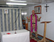 Chaplain Andrew Martlew in his chapel.  The room is small and the furniture seems almost makeshift, with benches, a rug on the floor, a crucifix on the wall and a curtain behind the small altar to divide the room in two