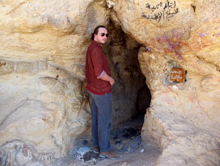 Nicholas Buxton at a small cave entrance cut in the striated yellow and beige stone.  Writing in a non-Western script is visible next to the doorway