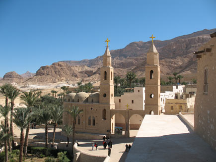 Saint Antony's monastery, a collection of warm-coloured stone buildings with small, arched windows.  Two steeples topped by Christian crosses stand either side of the double arch entrance.  Palm trees grow in small walled patches of green space outside the walls