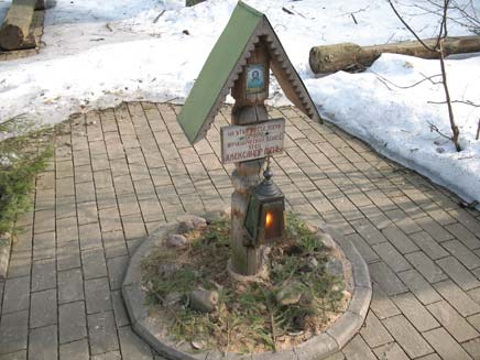 Memorial to Father Alexander: a small wooden pillar set in the middle of a paved path, surrounded by snow.  The display has a tiny roof to protect an image of Jesus and a plaque bearing Alexander's name from the elements. A lighted lantern is hanging underneath the plaque