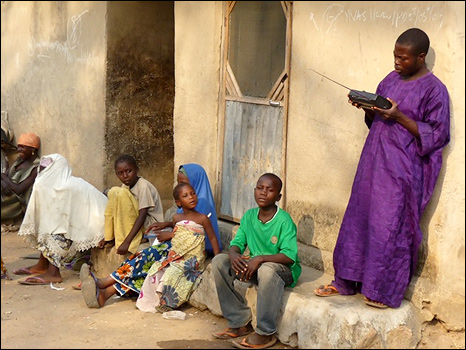 A man holds a radio as women and children sit at the side of the road