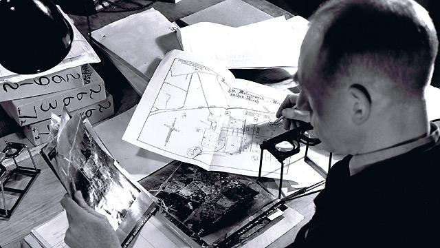 An RAF Flight Lieutenant Photo Interpreter busy with the tools of his trade - aerial photographs, stereoscopes and the plans of a target, in 1944. 