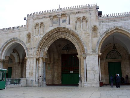 Facade of the Al-Aqsa mosque, a stone building on square lines with three arched doorways.  Visitors stand around talking and security lights are visible on the corners of the roof