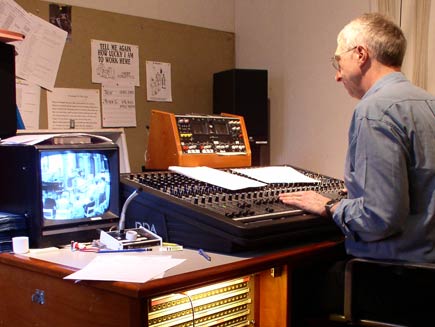 Looking over the shoulder of the sound engineer, who is balancing sound levels using the console in front of him.  A monitor on the desk shows the scene in the main room of the church.  On the far wall, a noticeboard displays a few clippings and cartoons, including one with the caption 'Tell me again how lucky I am to work here'