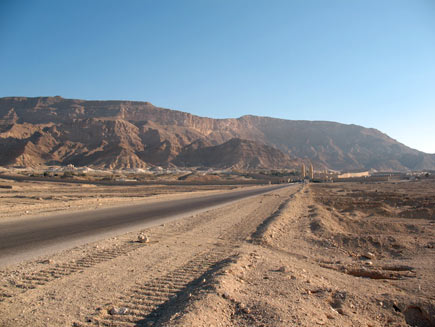 A road through the Egyptian desert.  Tyre marks can be seen in the sand on the tarmac.  In the distance a collection of low, light-coloured stone buildings is dwarfed by the red-brown mountains behind