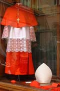 Vestments of a cardinal: a dummy wearing a red cassock, white rochet shirt trimmed with lace, red chimere robe and apostolical cross, with red zucchetto skullcap, white mitre and red stole at its feet, on display in a shop window