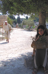 Galilee. A girl sits in the shade of a tree while a man walking with a staff goes past