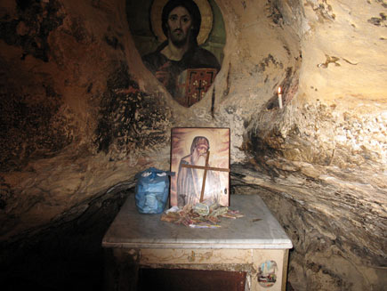 Inside the cave a small table serves as altar, with a plastic bag of papers, a pile of monetary donations, a painting of Saint Antony and a simple wooden cross.  On the wall behind, seeming to be painted directly on the stone, is a larger picture of Christ.  A small white candle burns in a niche in the rock