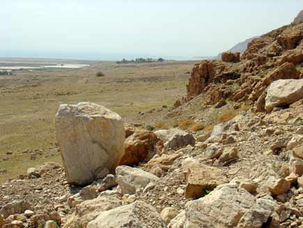 Loose scree and boulders on a hillside, with a tiny settlement visible in the distance and the shore of the Dead Sea visible on the left