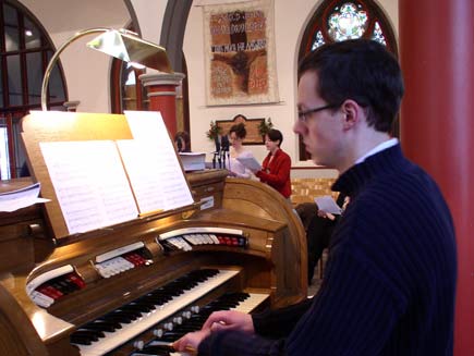 Greg Morris the organist sitting at the console of a small Conacher organ, with the choir members nearby in the background