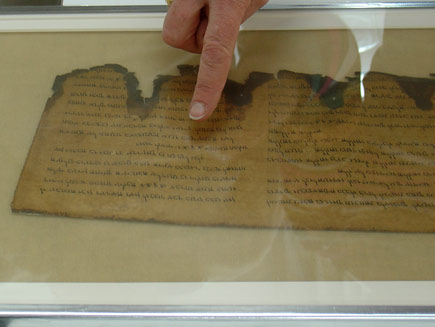 Pointing to a scroll that is preserved under glass. This scroll looks almost complete, with just the bottom edge showing jagged damage and discolouration