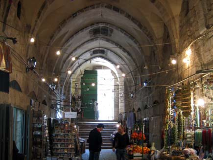Market stalls are set up in a covered alleyway with a high, arched ceiling.  An arched doorway in the background lets some light in, and electric lights are strung across the alley