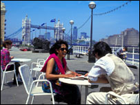 Casal aproveita o sol em café à beira do rio Tâmisa, tendo ao fundo a ponte da Torre de Londres