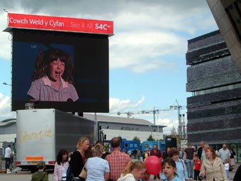 Ymwelwyr Eisteddfod yr Urdd yn gwylio'r cystadlu o'r llwyfan ar sgrin fawr y tu allan i Ganolfan Mileniwm Cymru